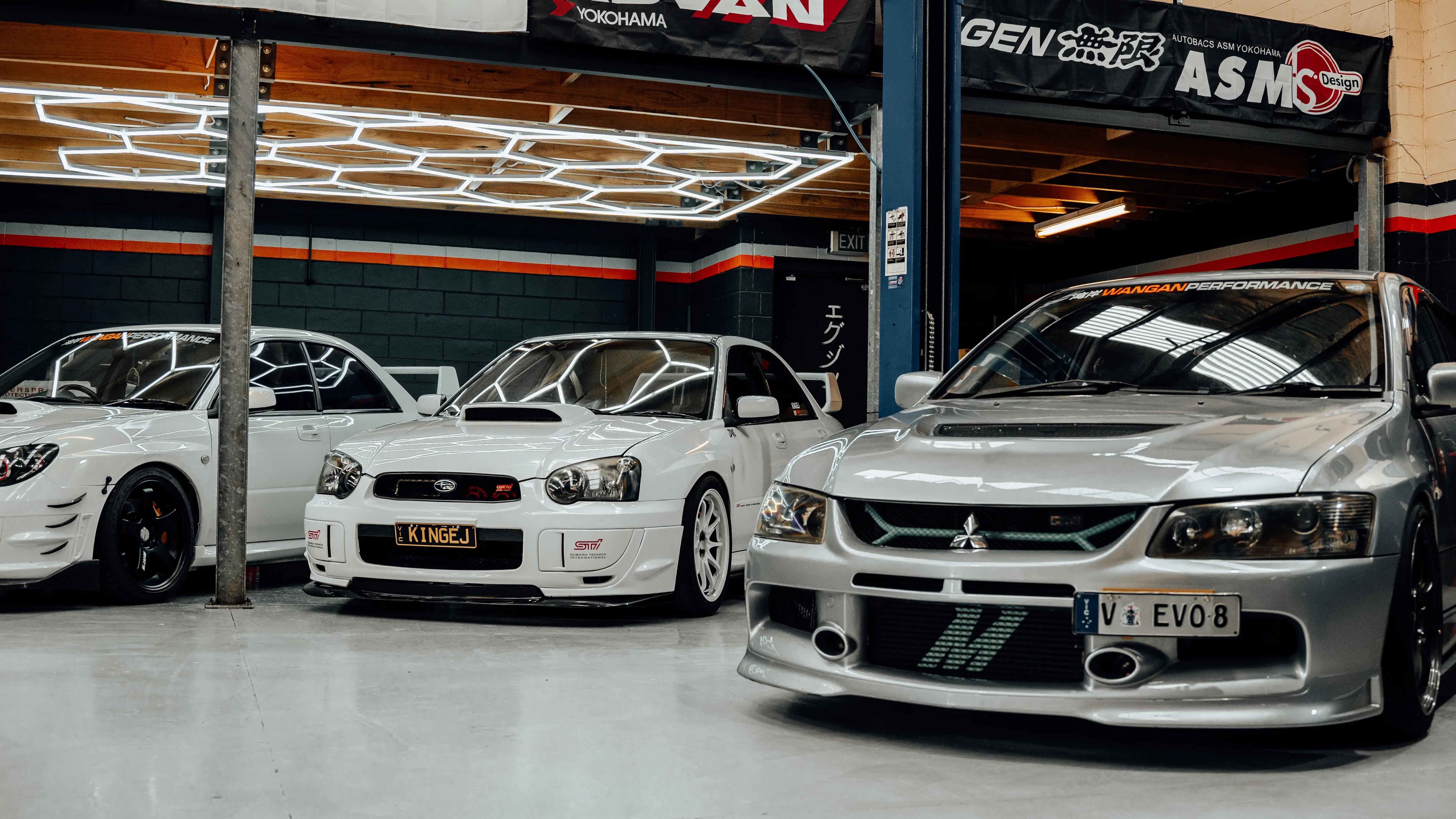 Subaru STI and Mitsubishi Evo under honeycomb workshop lights at Wangan Performance